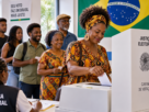 Eleitores brasileiros, majoritariamente negros, participam das eleições em uma seção eleitoral, com uma mulher negra votando e outras pessoas aguardando na fila.